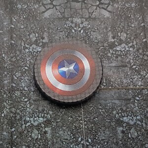 May include: A red, white, and blue Captain America shield with a silver star in the center. The shield is lying on a gray, textured surface.