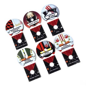 May include: A collection of Star Wars character tokens. Each token features a character's name, such as "Finn" and "Luke Skywalker", along with a stylized image of their helmet or face. The tokens are red, black, white, and green.