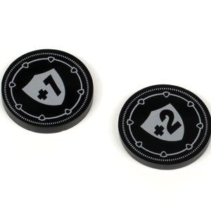 May include: Two black game tokens with white shield and number graphics. One token has a "1" and the other has a "2" with a plus sign. The tokens have a dotted border and are on a white background.