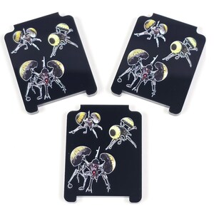 May include: Three black rectangular coasters with rounded corners, each featuring a repeating illustration of a mechanical insect-like creature with yellow and red accents. The coasters are arranged on a white surface.