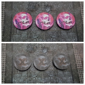 Könnte beinhalten: Drei rosafarbene und violette schädelförmige Spielsteine mit einem schwarzen X auf jedem. Drei graue Spielsteine mit einem schwarzen X auf jedem.