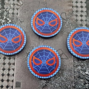 Puede incluir: Cuatro fichas redondas azules y rojas con una telaraña blanca y ojos rojos en el centro. Las fichas tienen un borde blanco punteado.