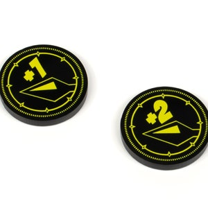 May include: Two black and yellow game tokens with a plus sign and a number. The first token has a plus sign and the number 1. The second token has a plus sign and the number 2.