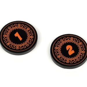May include: Two black circular game pieces with orange numbers, 1 and 2, on a white background.
