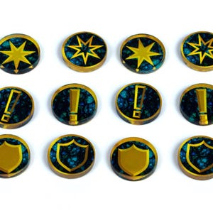 Puede incluir: Doce fichas de juego redondas con bordes dorados y símbolos. Las fichas presentan un fondo turquesa con diseños dorados de estrellas, signos de exclamación y escudos. Las fichas están dispuestas en tres filas de cuatro.