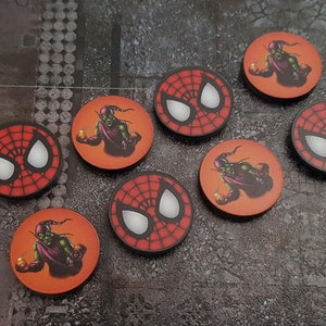 Può includere: Gettoni di gioco circolari con la maschera di Spider-Man e Goblin. I gettoni sono rossi con ragnatele nere e occhi bianchi, e arancioni con Goblin. I gettoni sono disposti su una superficie grigia.