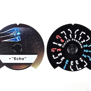 Könnte beinhalten: Zwei schwarze, kreisförmige Spielmarken. Eines zeigt eine blau-weiße Raumschiffgrafik und den Text ". Echo". Das andere hat weiße und farbige Zahlen und Pfeile. Die Marken sind für ein Brettspiel.