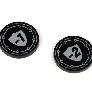 Puede incluir: Dos fichas de juego de plástico negro con un diseño de escudo plateado. El escudo tiene un número, una ficha tiene el número 1 y la otra el número 2.
