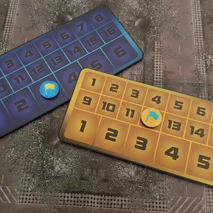 May include: Two game boards with numbers 1 to 16. The board on the left is blue with a blue flag marker on space 12. The board on the right is yellow with a yellow flag marker on space 11.