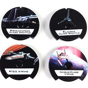 May include: Four circular black game tokens featuring Star Wars starships. The tokens are labeled Sheathipede Class Shuttle, Lambda Class Shuttle, T65 X-Wing, and Nimbus-Class V-Wing, each with a central circular cutout and notched edges.