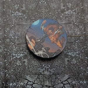 May include: A round, black and white token featuring a colourful image of a superhero in mid-air, with a blue background and a moon in the top left corner.