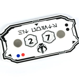 May include: A white plastic game token with black markings and a Mandalorian symbol. The token has two numbers, "2" and "7", and the text "E1V D01M1V".