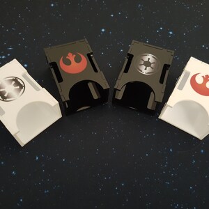 Könnte beinhalten: Vier Acryl-Würfeltürme, zwei schwarz und zwei weiß. Jeder Turm zeigt ein anderes Star Wars-Logo: das Symbol der Rebellenallianz in Rot, das imperiale Symbol in Silber und das Symbol der Rebellenallianz erneut in Rot. Die Türme sind in einer Reihe auf einem schwarzen Hintergrund mit weißen Punkten angeordnet.