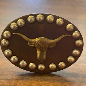May include: A brown leather belt buckle with a gold-toned steer head design. The buckle is surrounded by a row of silver-toned studs.