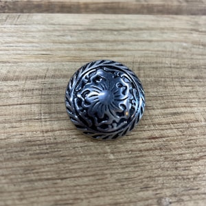 May include: A silver-toned metal button with a decorative floral design. The button has a raised, textured edge.