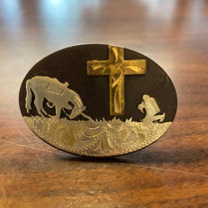 May include: Oval-shaped belt buckle with a dark background and gold and silver accents. The design features a gold cross, a horse, and a person kneeling in prayer. The bottom edge has a decorative silver pattern.