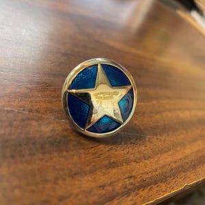 May include: A silver-toned star-shaped button with a blue enamel center. The star has a raised design and the button has a smooth, rounded edge.