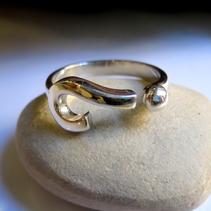 May include: A silver ring with a unique, abstract design. The ring features a curved, open shape with a small, round bead at the end.