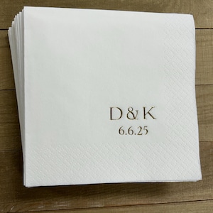 May include: A stack of white paper napkins with a textured surface. The napkins are printed with the initials "D&K" and the date "6.6.25" in a gold color. The napkins are likely for a wedding or special event.