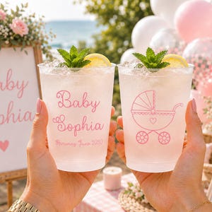 May include: Two clear plastic cups filled with ice, a lemon slice, and a mint sprig. One cup features the pink text Baby Sophia and Arriving June 2030, while the other displays a pink line drawing of a baby carriage with a small heart in the center.