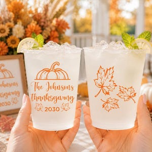 May include: Two clear plastic cups filled with ice and a light-colored beverage, garnished with lemon slices and mint. The cups feature orange graphics of a pumpkin, maple leaves, and the text The Johnsons thanksgiving 2030 printed on them.