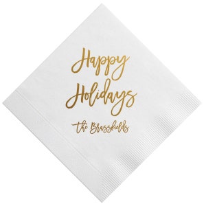 May include: White paper napkin with gold foil lettering that reads "Happy Holidays" and "The Brassfields".
