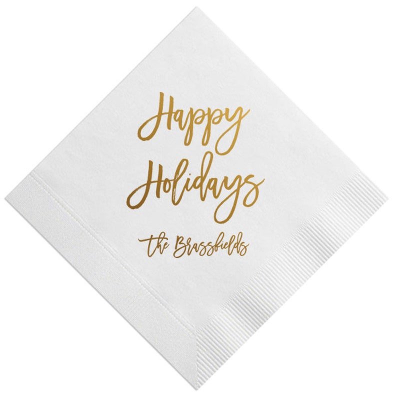 May include: White paper napkin with gold foil lettering that reads "Happy Holidays" and "The Brassfields".