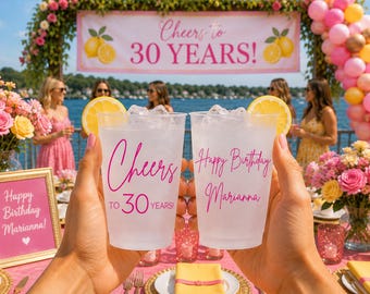 May include: Two clear plastic cups filled with ice and a lemon slice on the rim. The left cup features pink text reading Cheers TO 30 YEARS! and the right cup features pink text reading Happy Birthday Marianna in a cursive script.