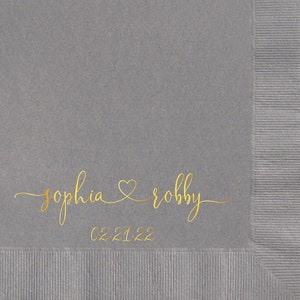 May include: Gray paper napkin with gold lettering that reads "sophia & robby" with a heart symbol between the names and the date "02.21.22" below.