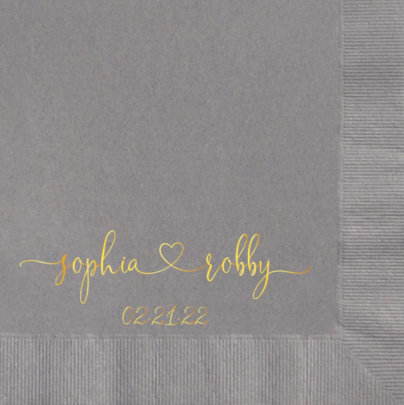 May include: Gray paper napkin with gold lettering that reads "sophia & robby" with a heart symbol between the names and the date "02.21.22" below.