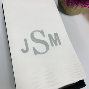 May include: A white cloth napkin with a silver monogram of the letters "JSM" embroidered on it. The napkin is folded and sitting on a black surface.