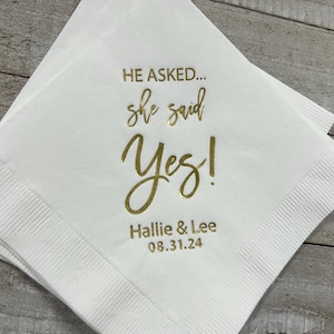 May include: White paper napkin with gold foil lettering that reads "He asked... she said Yes! Hallie & Lee 08.31.24".