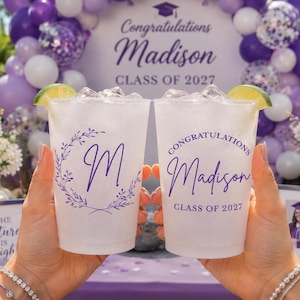 May include: Two clear plastic cups filled with a beverage and a lime wedge. One cup features a purple letter M inside a leafy wreath. The other cup displays the text Congratulations Madison CLASS OF 2027 in purple ink on the front surface.