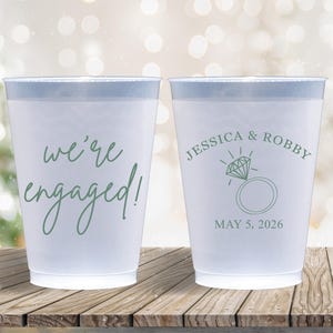 May include: Two white plastic cups with green text. One cup says "We're engaged!" and the other says "Jessica & Robby" with a diamond ring and "May 5, 2026".