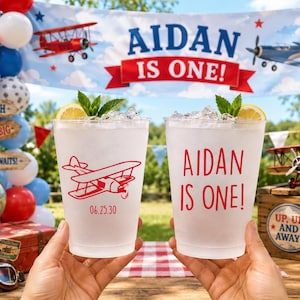 May include: Two clear plastic cups filled with ice, lemon slices, and mint. One cup features a red airplane graphic and the date 06.25.30, while the other displays the red text AIDAN IS ONE! in a bold, sans-serif font.