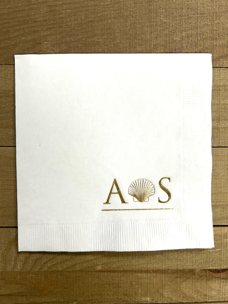 May include: White paper napkin with gold foil lettering "A  S" with a seashell graphic between the letters.