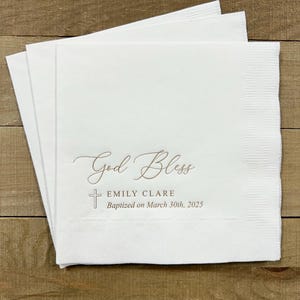May include: White paper napkins with the text "God Bless" in cursive script, a small cross, and the text "Emily Clare Baptized on March 30th, 2025".