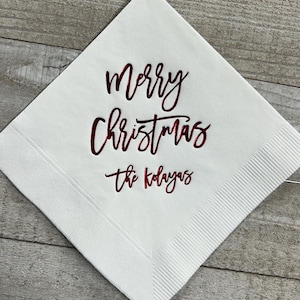 May include: White paper napkin with the words "Merry Christmas" in red cursive lettering and "The Kolayas" in smaller red cursive lettering.