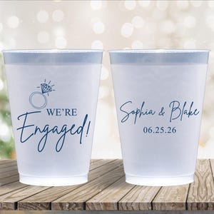 May include: Two white plastic cups with blue text. One cup says "We're Engaged!" with a diamond ring icon. The other cup says "Sophia & Blake 06.25.26".