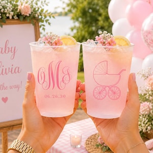May include: Two clear plastic cups filled with a pink beverage, ice, a lemon slice, and small pink flowers. One cup features a pink monogram with the date 06.26.30, while the other displays a pink line-art illustration of a baby carriage.