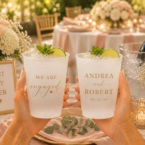 May include: Two frosted plastic cups filled with ice, lime wedges, and mint leaves. One cup features gold text reading WE ARE engaged with a heart, and the other displays ANDREA and ROBERT 06.26.30 in gold lettering against a translucent surface.
