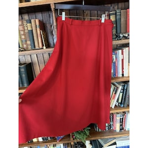 May include: A vibrant red skirt hanging on a wooden hanger. The skirt has a classic A-line silhouette, a waistband with belt loops, and a subtle front pleat. The fabric appears to be a smooth, solid color, suitable for various occasions.