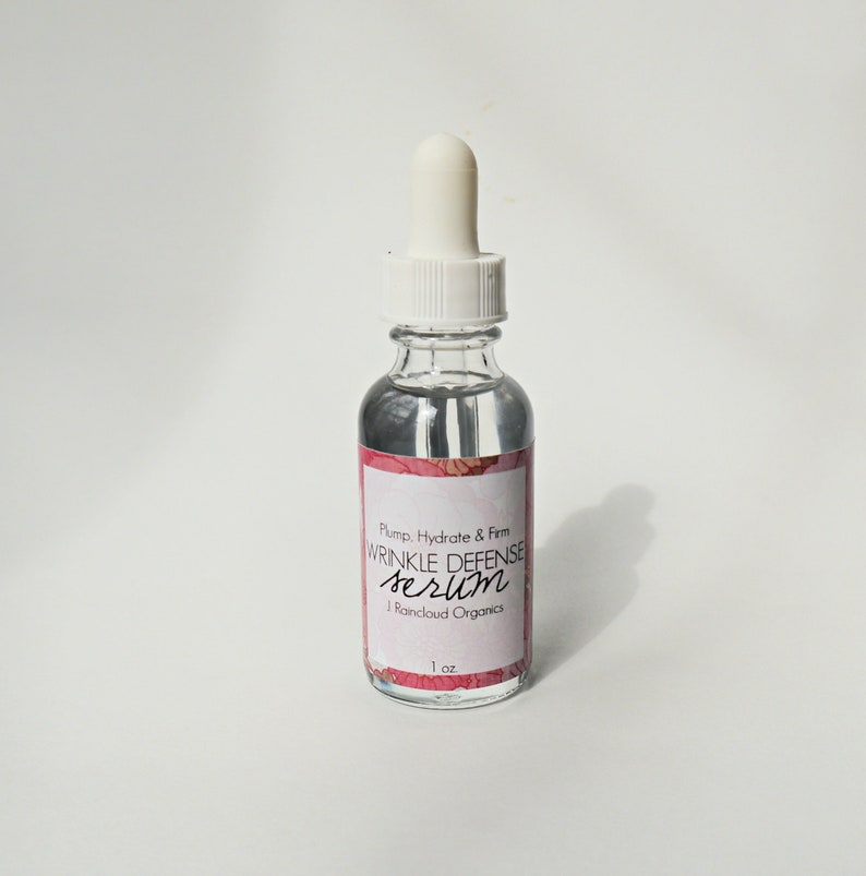 May include: A clear glass bottle with a white dropper top and a label that reads 'Plump, Hydrate & Firm Wrinkle Defense Serum J. Rainaud Organics 1 oz.'