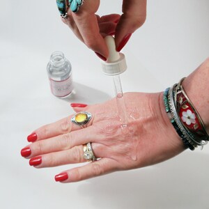 Puede incluir: Una mano con esmalte de u&ntilde;as rojo est&aacute; recibiendo gotas de l&iacute;quido transparente de un frasco gotero. La mano lleva varias pulseras de plata, una con un dise&ntilde;o de flor roja y blanca. La persona tambi&eacute;n lleva un anillo con una piedra amarilla.