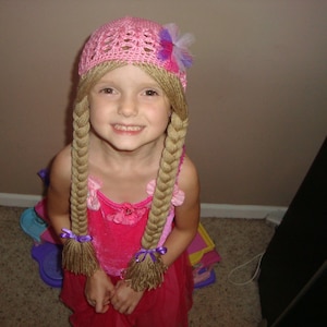 May include: A young girl wearing a pink crocheted hat with a purple flower and long blonde braids. She is wearing a pink dress with a floral pattern and a purple ribbon in her hair.