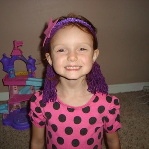May include: A young girl with purple yarn hair extensions and a pink and purple headband smiles at the camera. She is wearing a pink and black polka dot dress.