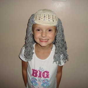 May include: A young person wearing a white crocheted hat with gray yarn strands hanging down, resembling hair. They are wearing a white t-shirt with the text "BIG SIS" in pink and blue.