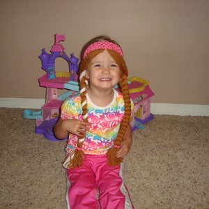 May include: A young girl with long brown hair wearing a pink headband and a colorful shirt is playing with a pink and purple toy castle.