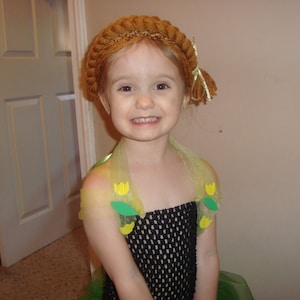 May include: A young girl wearing a brown crocheted hat with yellow flowers and a green and black dress with yellow flowers.