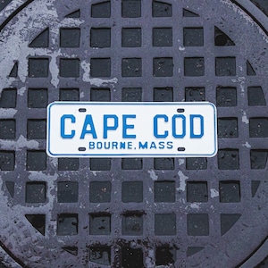 May include: A white metal sign with blue lettering that reads "CAPE COD BOURNE, MASS". The sign is placed on top of a gray metal sewer grate.